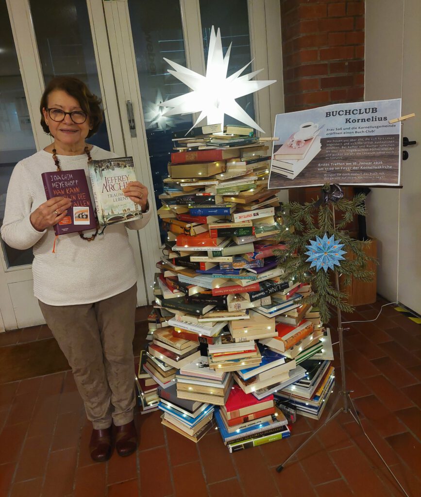 Eine Frau mit Büchern in den Händen steht neben einem Weihnachtsbaum ganz aus Büchern gemacht, samt Lichterkette und Stern an der Bücherstapelspitze. 